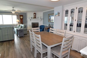 A dining area with a wooden table and white chairs, a blue armchair, a fireplace, and a clock on the wall. - 10% off 3-7 nights thru Oct. Incredible views! 3BR PH. Outdoor pool! Ocean Drive. (North Myrtle Beach)