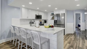A modern kitchen with white cabinetry, a central island with bar stools, a stainless steel refrigerator, and a microwave. - Oceanfront Penthouse with Sweeping Atlantic Views | Wild Dunes | Isle of Palms (Isle of Palms)