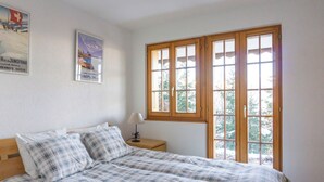 Ein Schlafzimmer mit einem Bett, einem Nachttisch, einer Lampe und einem Fenster mit Blick auf Bäume.