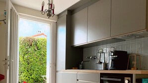 A kitchen with a built-in oven, a coffee maker, and a view of a garden through an open door. - Seaside Holiday Home: Spacious, Welcoming, Garden, Wi-Fi (Pornic)