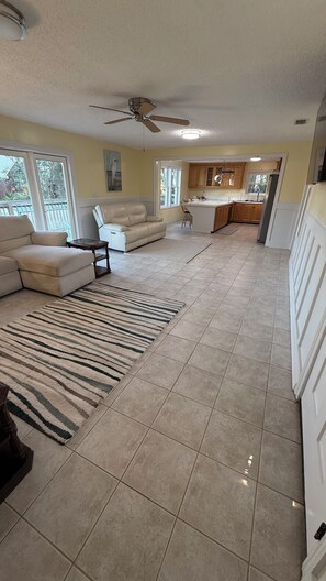 A spacious living room with a beige sofa, a coffee table, and a striped rug. There is a kitchen area with wooden cabinets and a ceiling fan.