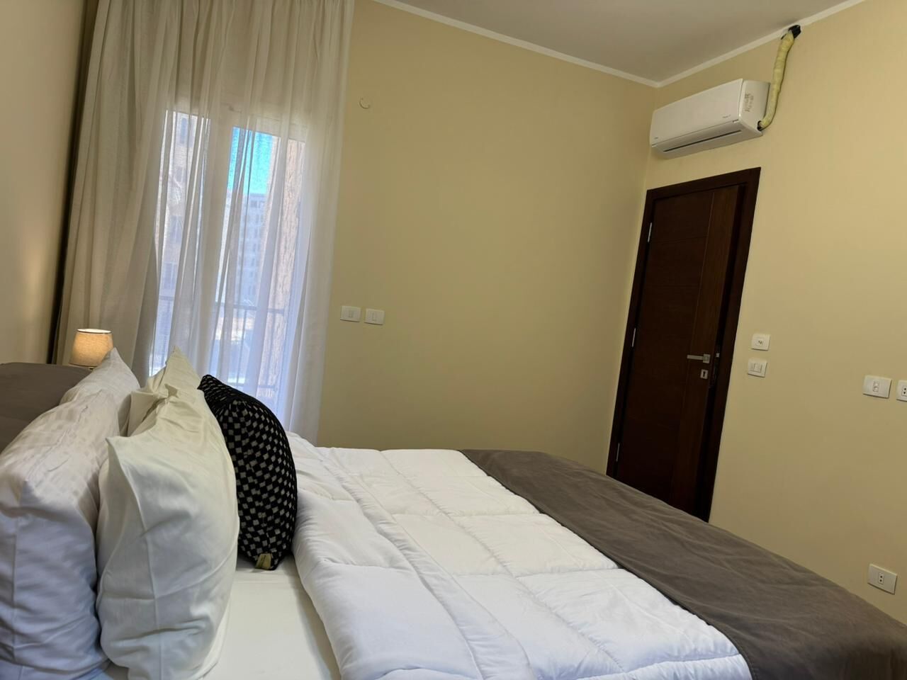 A hotel room with a bed, a window with curtains, an air conditioning unit, and a door.