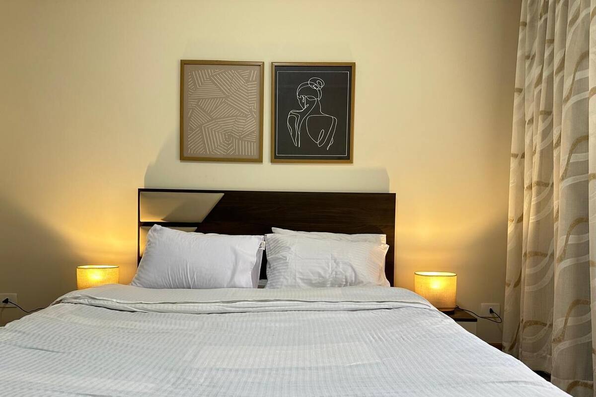 A neatly made bed with white bedding, two wall-mounted lamps, and two framed artworks above it.