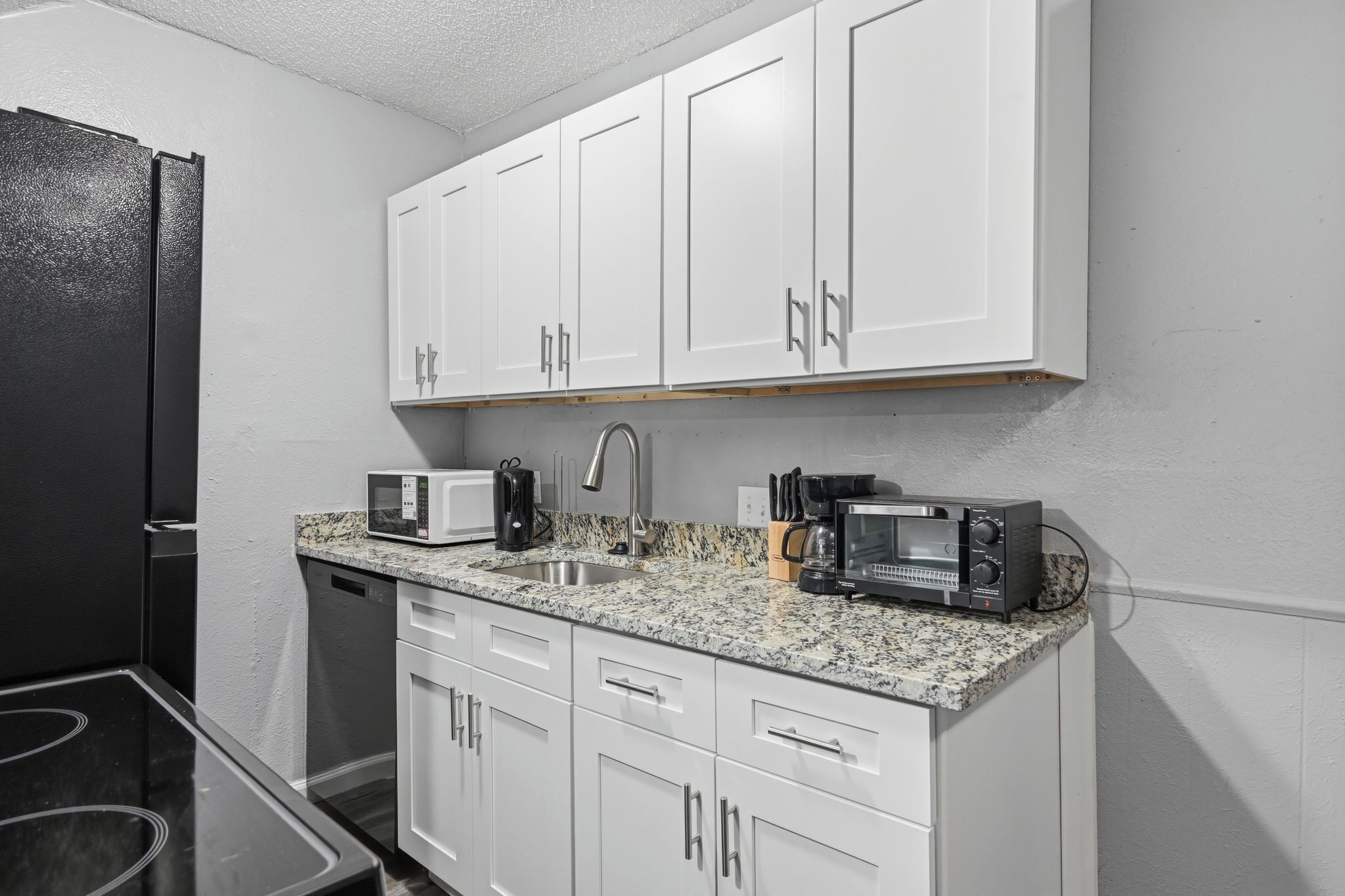 A kitchen with white cabinets, a black refrigerator, a granite countertop, a microwave, a coffee maker, and a toaster.