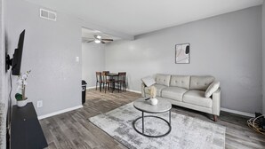 Uma sala de estar com sofá, mesa de centro, mesa de jantar com cadeiras e uma TV fixada na parede.