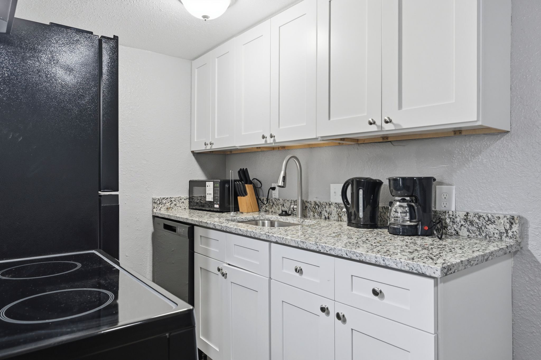 Una cocina con armarios blancos, un refrigerador negro, un microondas, una cafetera y un lavamanos.