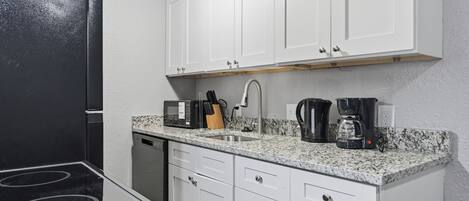 Una cocina con armarios blancos, un refrigerador negro, un microondas, una cafetera y un lavamanos.
