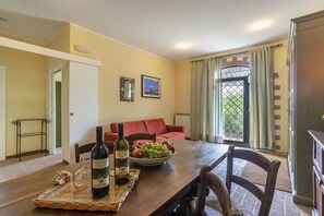 A dining area with a wooden table, two bottles of wine, a bowl of fruit, a red sofa, and a view through a window with curtains. - Nice room for 4 people with WIFI, A/C, pool, TV, veranda and pets allowed, close to Perugia (Agello)