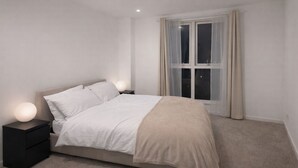 A bedroom with a bed, bedside tables, a window with curtains, and a carpeted floor. - Round The Clock Bookings - Wharf 1ne (Barking)