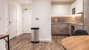 A modern kitchen with a built-in oven, a stainless steel trash bin, and a dining area with a round table. - Round The Clock Bookings - Wharf 1ne (Barking)