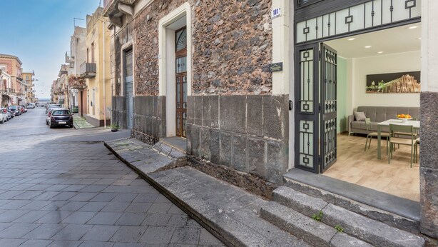 A street view with a building featuring a stone facade, a gated entrance, and a dining area visible through the window. - 'Vitali Home' - Apartment in the Heart of Catania (Catania)