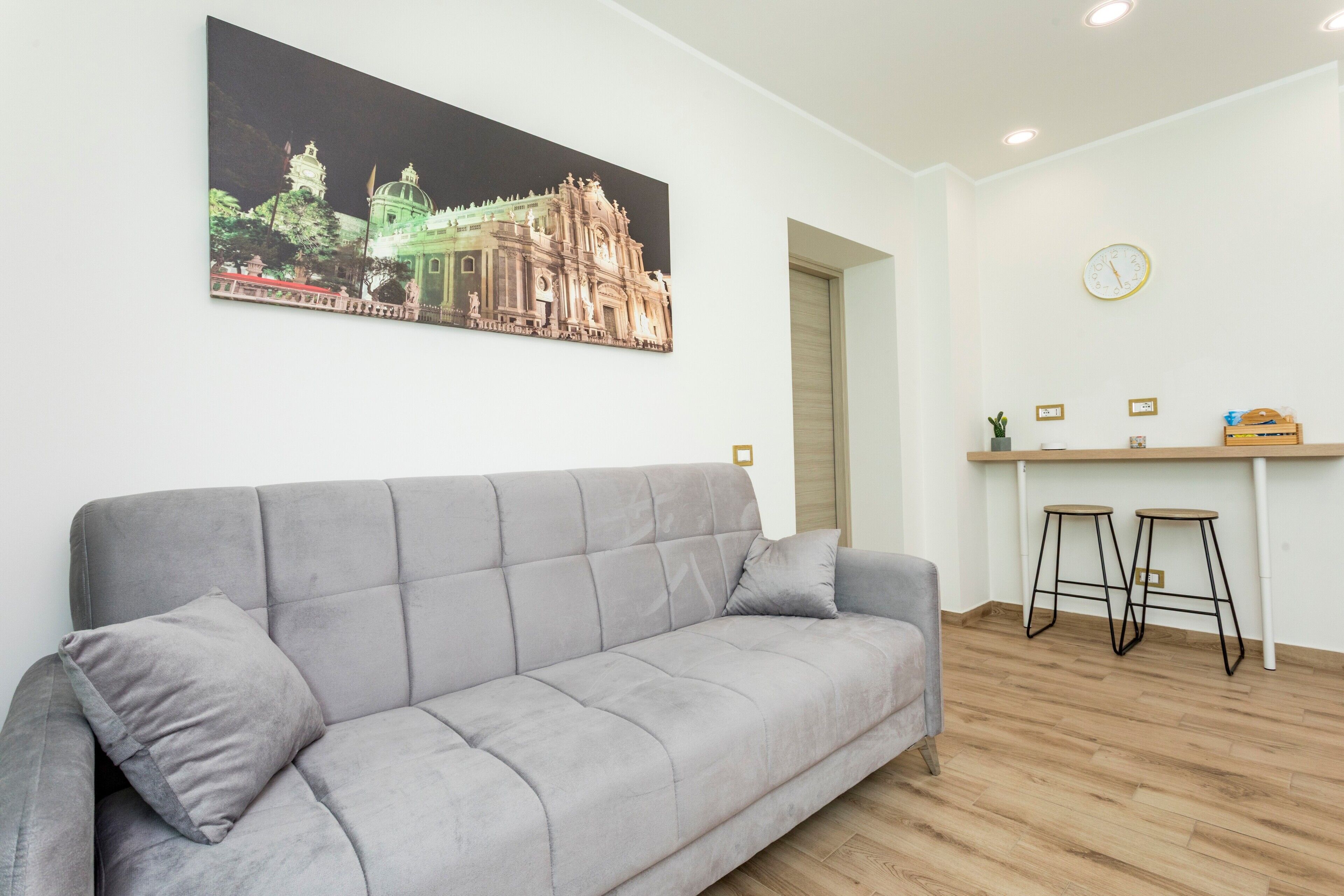 Ein modernes Wohnzimmer mit einer grauen Couch, einem Essbereich mit Hocker, einer an der Wand befestigten Uhr und einem gerahmten Bild eines Gebäudes.