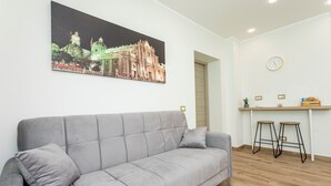 A modern living room with a grey sofa, a dining area with stools, a wall-mounted clock, and a framed picture of a building. - 'Vitali Home' - Apartment in the Heart of Catania (Catania)
