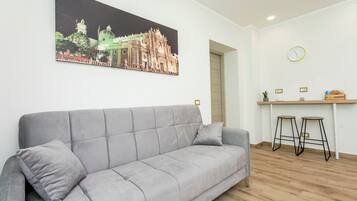 Ein modernes Wohnzimmer mit einer grauen Couch, einem Essbereich mit Hocker, einer an der Wand befestigten Uhr und einem gerahmten Bild eines Gebäudes.