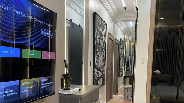 Een moderne hotelkamer met een flatscreen-tv waarop slimme functies worden getoond, een bureau met stoel, een bed en een hal met een spiegel en kunstwerken.