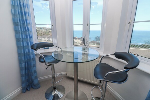 A dining area with a round glass table and two black chairs, a blue curtain, and a view of the sea through large windows. - Seaside Retreat (St Ives)