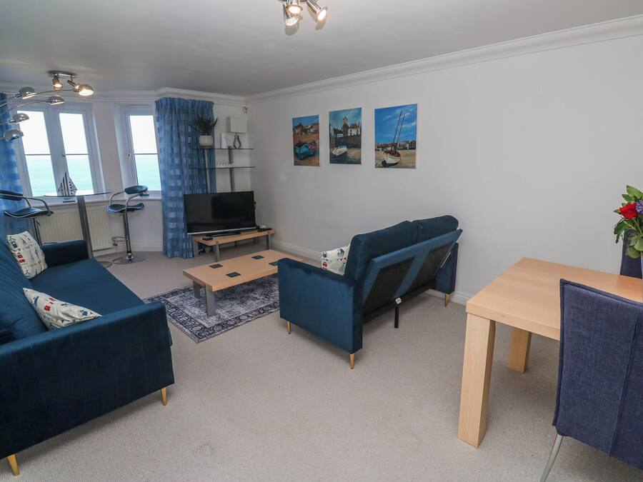 A living room with a sofa, a TV, a dining table, and a chair. There are three windows with curtains, a plant, and artwork on the wall.