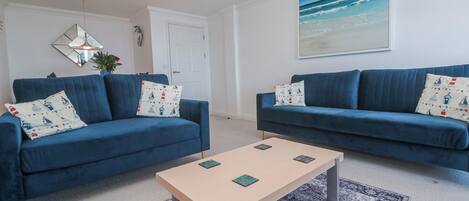 Una sala de estar con dos sofás azules, una mesa de centro color beige, un tapete estampado, un cuadro de una playa y un espejo en la pared.