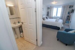 A bedroom with a bed, a blue armchair, a bookshelf, and a bathroom with a sink and mirror. - Seaside Retreat (St Ives)
