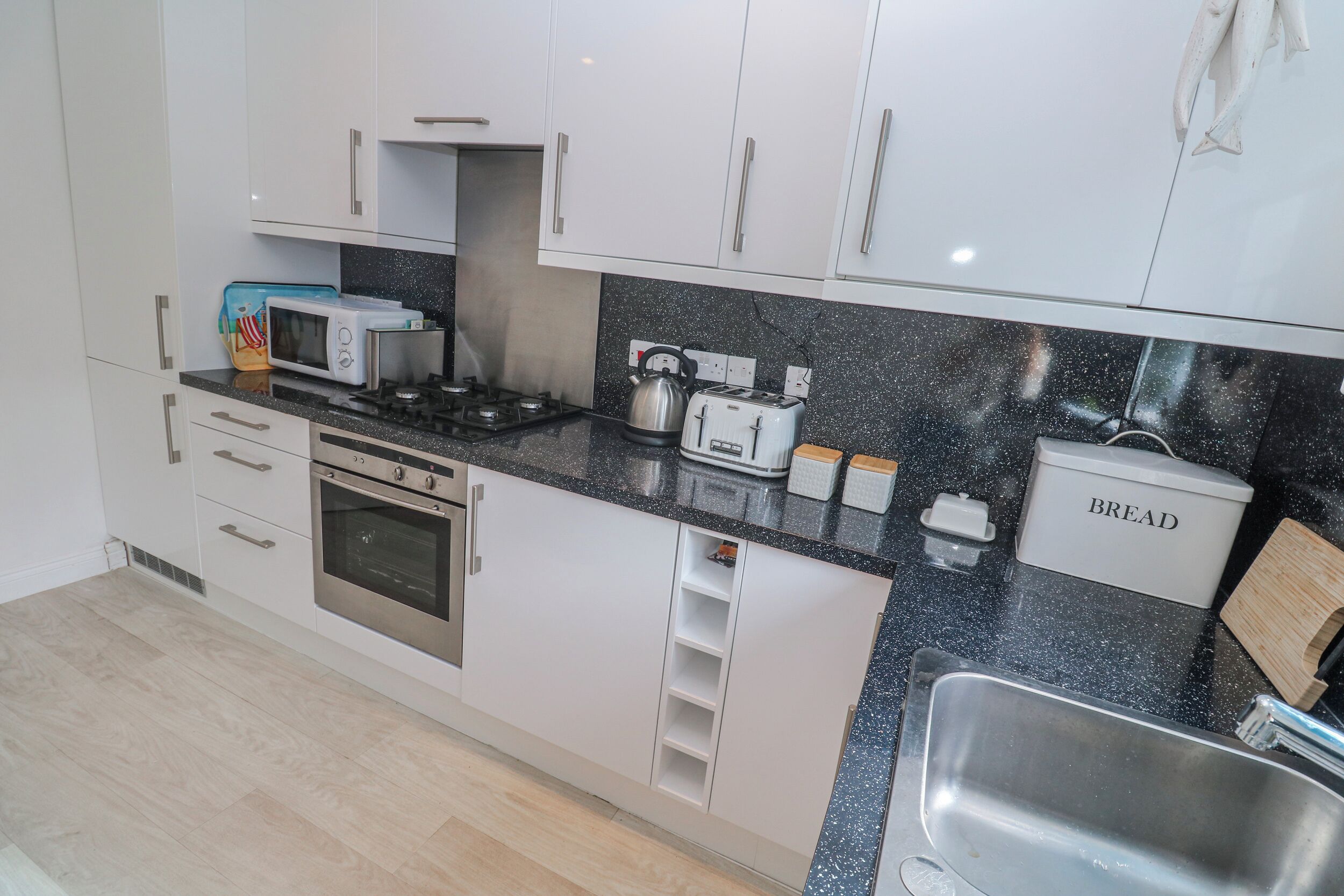 A modern kitchen with white cabinets, a stainless steel sink, a microwave, a kettle, a toaster, and a bread container.