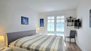 Un dormitorio con cama, lámparas de noche, una silla, televisor y ventana con vistas.