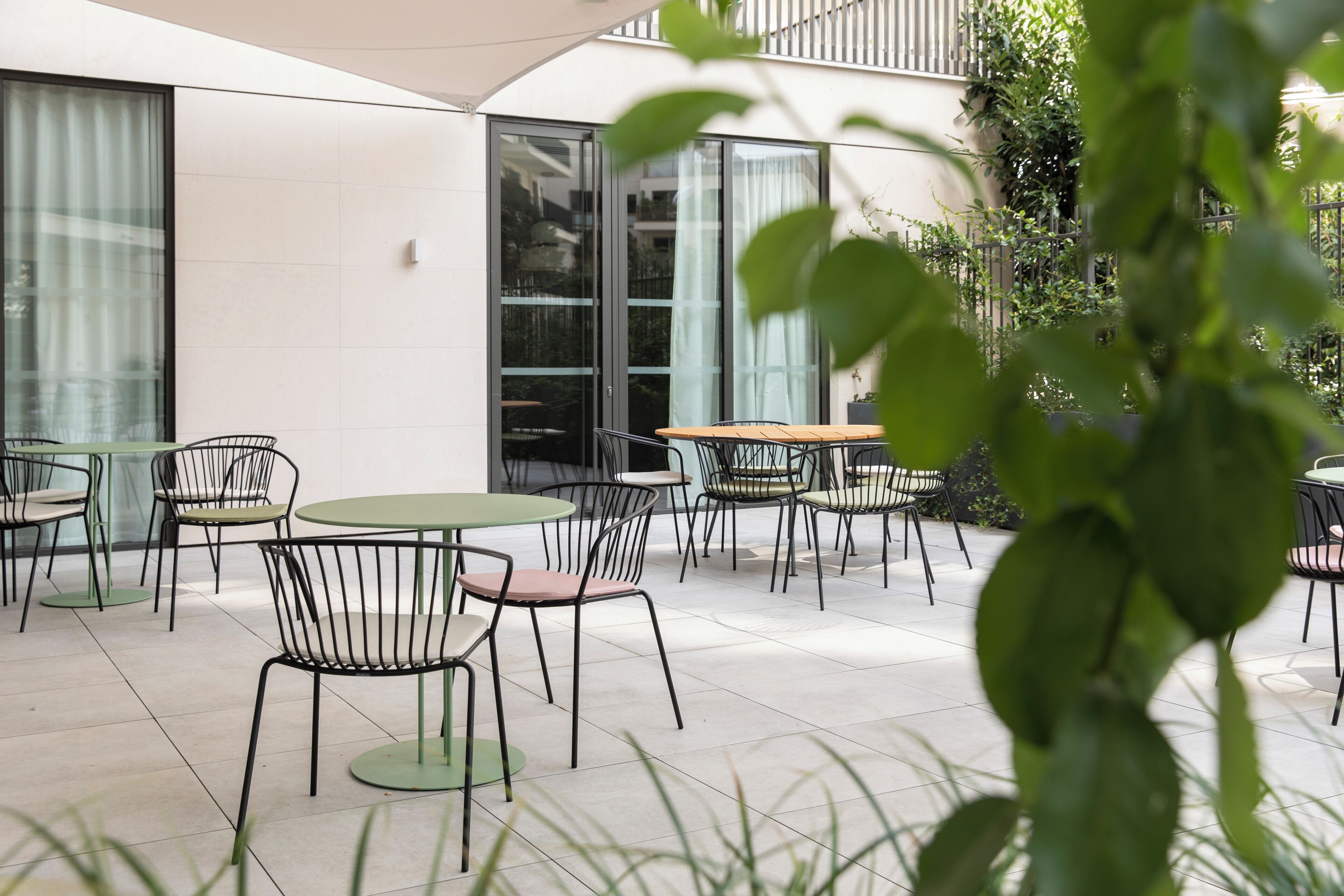 An outdoor patio area with round tables and chairs, a white building with large windows, and greenery in the foreground.