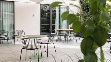 An outdoor patio area with round tables and chairs, a white building with large windows, and greenery in the foreground.