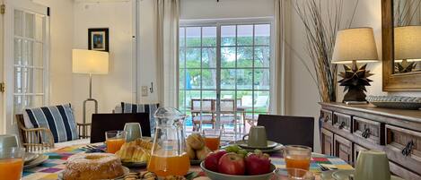Una mesa de desayuno con pasteles, frutas y bebidas, una ventana con vistas a la vegetación, y una lámpara al lado.