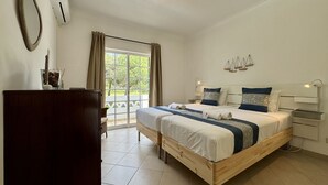 A bedroom with a wooden bed, a nightstand, a desk, a chair, a large window with curtains, and a ceiling light fixture. - Vilamoura Summer Villa 3 by Homing (Loulé)