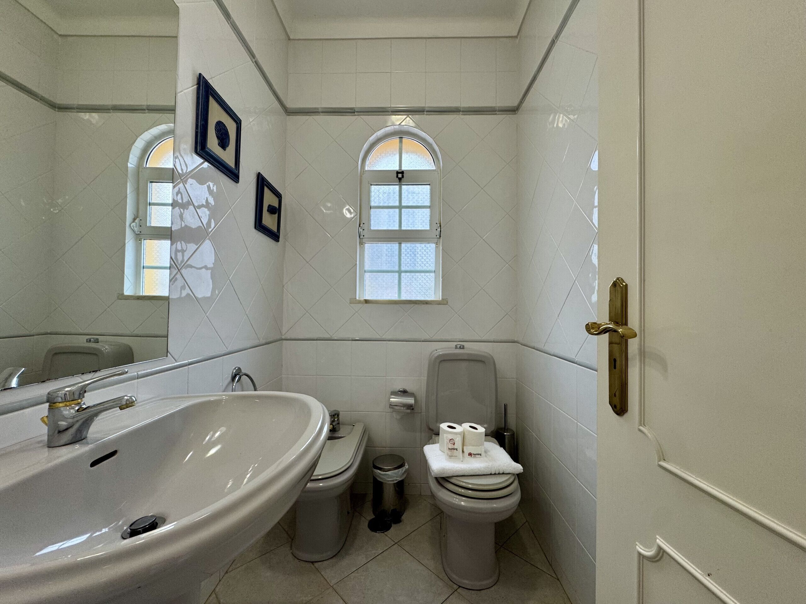 Ein Badezimmer mit Waschbecken, Toilette und Bidet, mit gefliesten Wänden und zwei kleinen Fenstern.