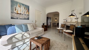 A living room with a sofa, coffee table, and a painting of sailboats. There is a dining area with chairs and a table. - Vilamoura Summer Villa 3 by Homing (Loulé)