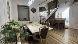 A modern dining area with a wooden floor, a white dining table, and chairs. There is a staircase with a black railing leading to an upper level. - Liz Ani Apartment (Abuja)