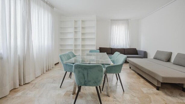 Een moderne woonkamer met een eettafel met glazen blad, blauwgroene stoelen, een grijze bank en een witte boekenkast.