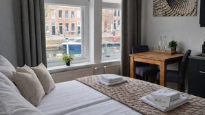 A hotel room with a bed, a table with chairs, a window with a view of buildings and a canal, and a welcome sign on the bed. - Studio Zeeland - central location - kitchen - North Sea beach 25 min (Goes)