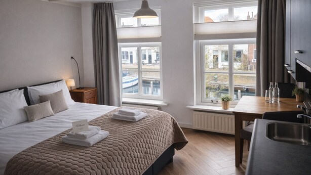 A hotel room with a bed, a desk, a view of buildings, and a window with curtains. - Studio Zeeland - central location - kitchen - North Sea beach 25 min (Goes)