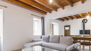 Ein modernes Wohnzimmer mit einer Couch, einem Glastisch, Holzbalken an der Decke, einem Esstisch mit Bänken und einem großen Spiegel.