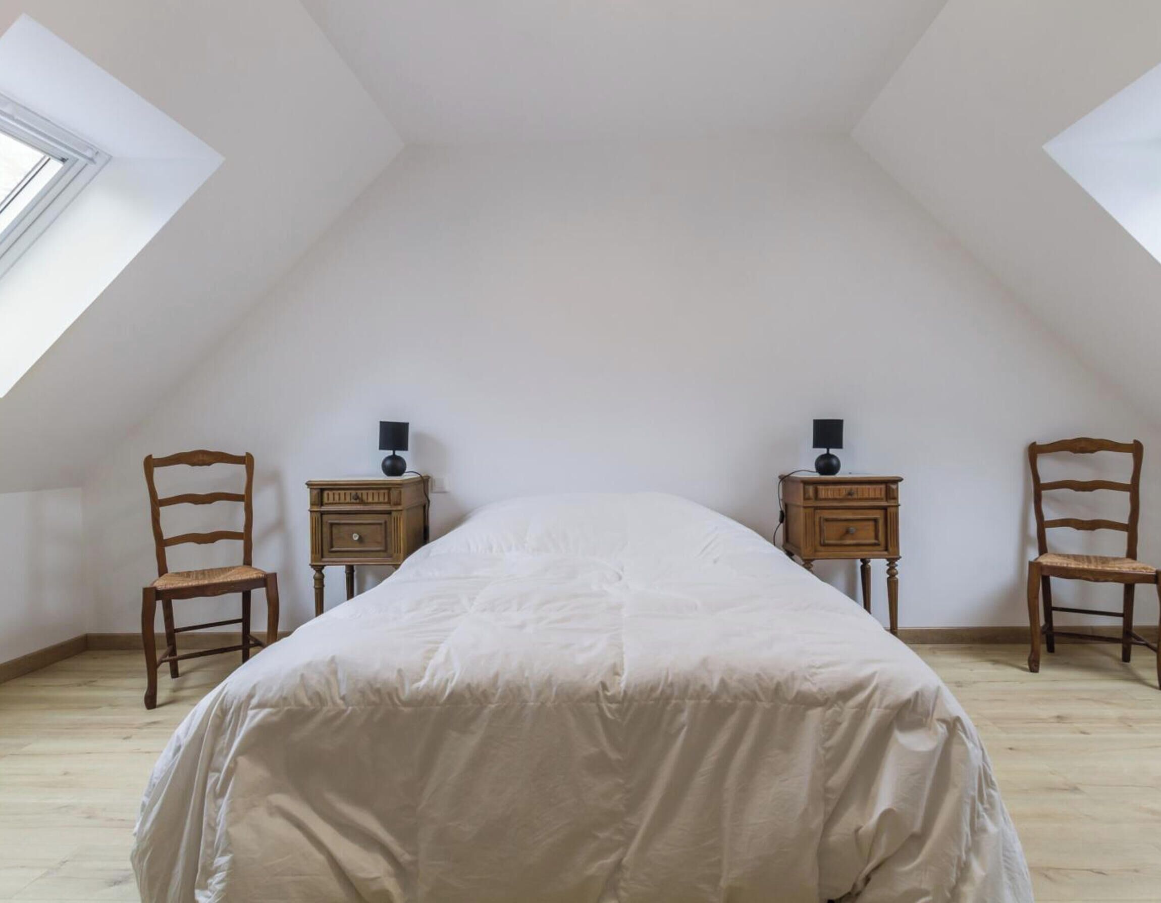 A high-ceilinged room with a sloped ceiling, a bed with a white duvet, two wooden nightstands with lamps, and two wooden chairs.
