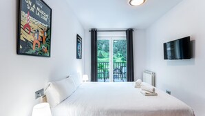 A hotel room with a bed, a TV, a window with a view of greenery, and a poster on the wall.