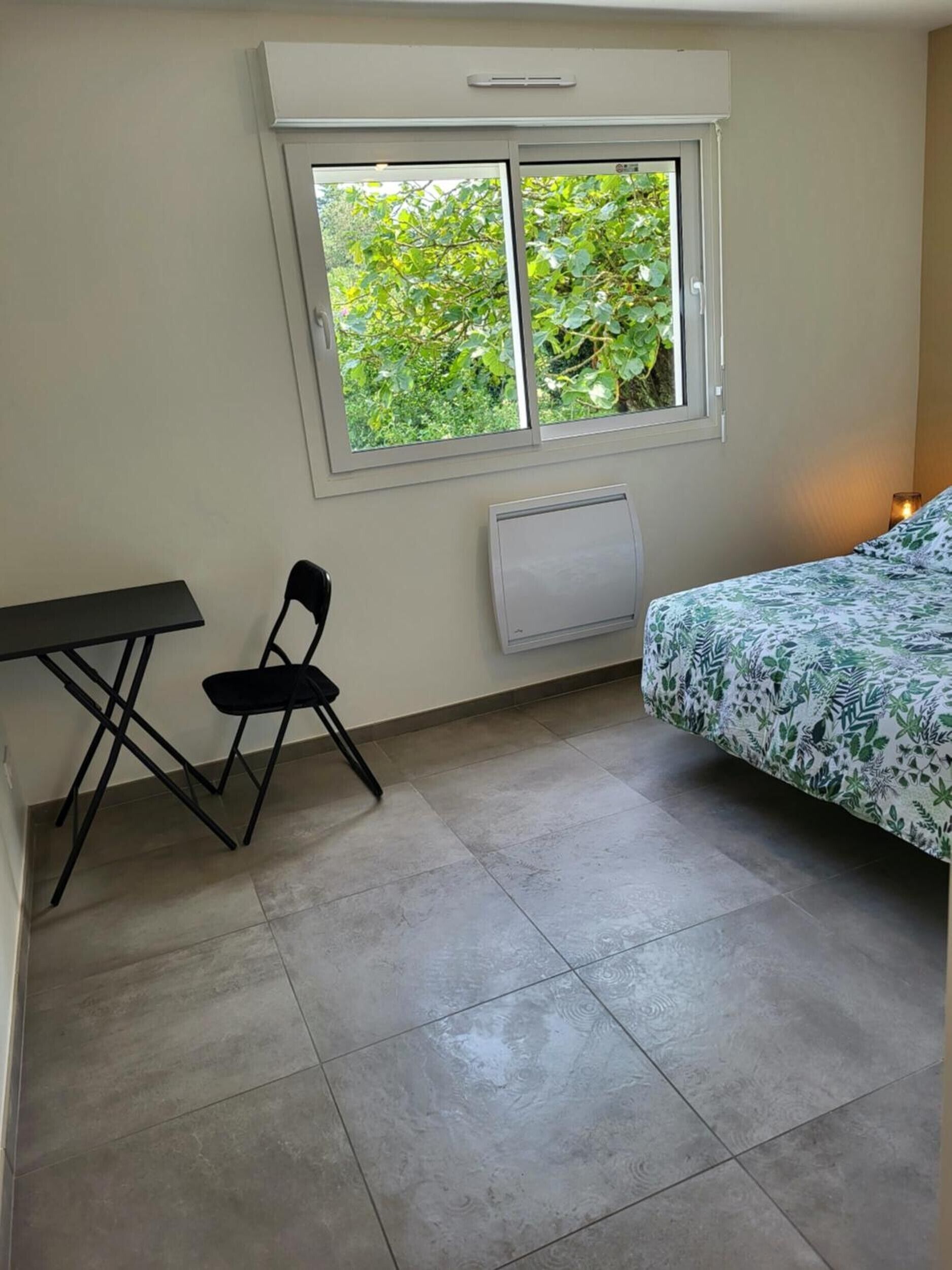 Et lille værelse med seng, bord, stol, et vindue med udsigt over grøn vegetation og en radiator.