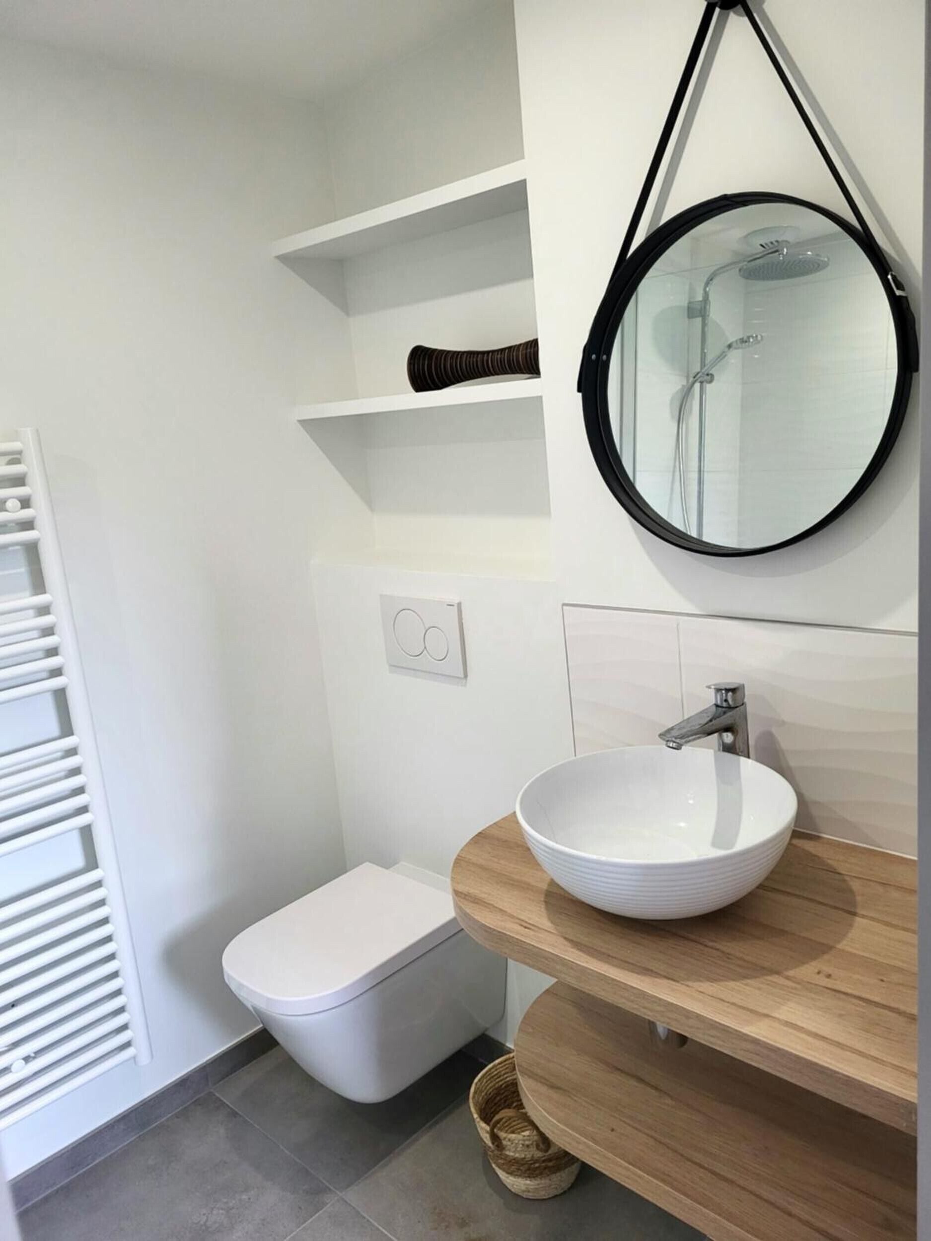 Une salle de bain moderne avec des toilettes, un miroir rond et un lavabo blanc posé sur un plan de travail en bois.