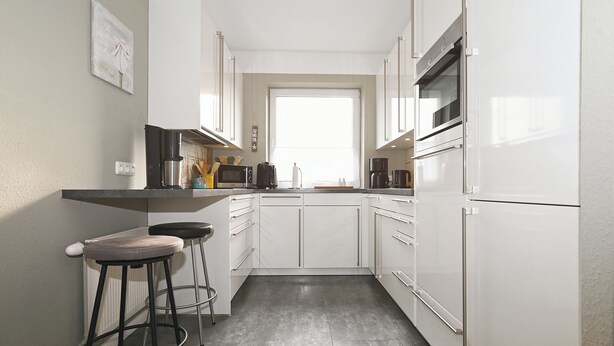 Eine kleine Küche mit weißen Schränken, einem Backofen, einer Mikrowelle und einer Kaffeemaschine. Zwei Barhocker und ein Fenster sorgen für natürliches Licht.