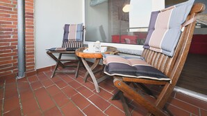 Ein hölzerner Schaukelstuhl mit gestreifter Kissenauflage, ein kleiner Holztisch mit Tasse und einer Schachtel, und ein Fenster mit Blick ins Innere.