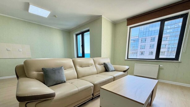 Una stanza con un divano beige, un tavolino bianco e ampie finestre che offrono vista sugli edifici.