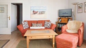 A living room with a sofa, ottoman, coffee table, television, bookshelf, and artwork. - Ostdeich Stübchen vacation apartment: Balcony & beach nearby (Büsum)