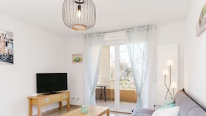 Una sala de estar con televisión, una mesa de centro de madera, un sofá con cojines, una lámpara colgante y un ventanal corredero que da a un balcón.