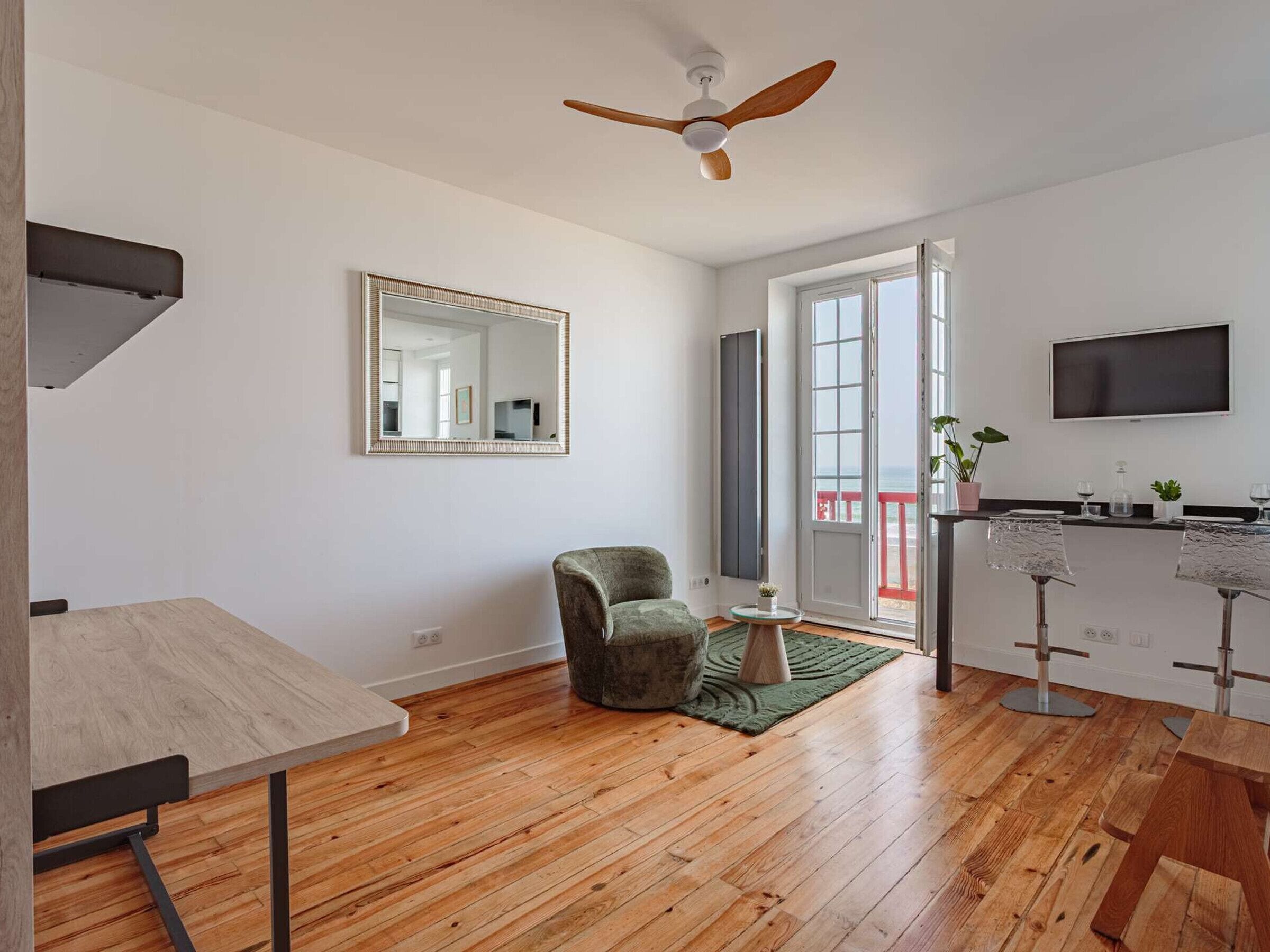 Een moderne woonkamer met houten vloer, een plafondventilator, een grote spiegel, een groene fauteuil, een klein rond tafeltje, en een keuken met een televisie en barstoelen.