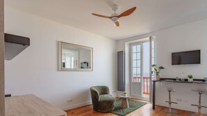 Ein modernes Wohnzimmer mit Holzboden, Deckenventilator, großem Spiegel, einem grünen Sessel, einem kleinen runden Tisch und einer Küchenzeile mit Fernseher und Barhockern.