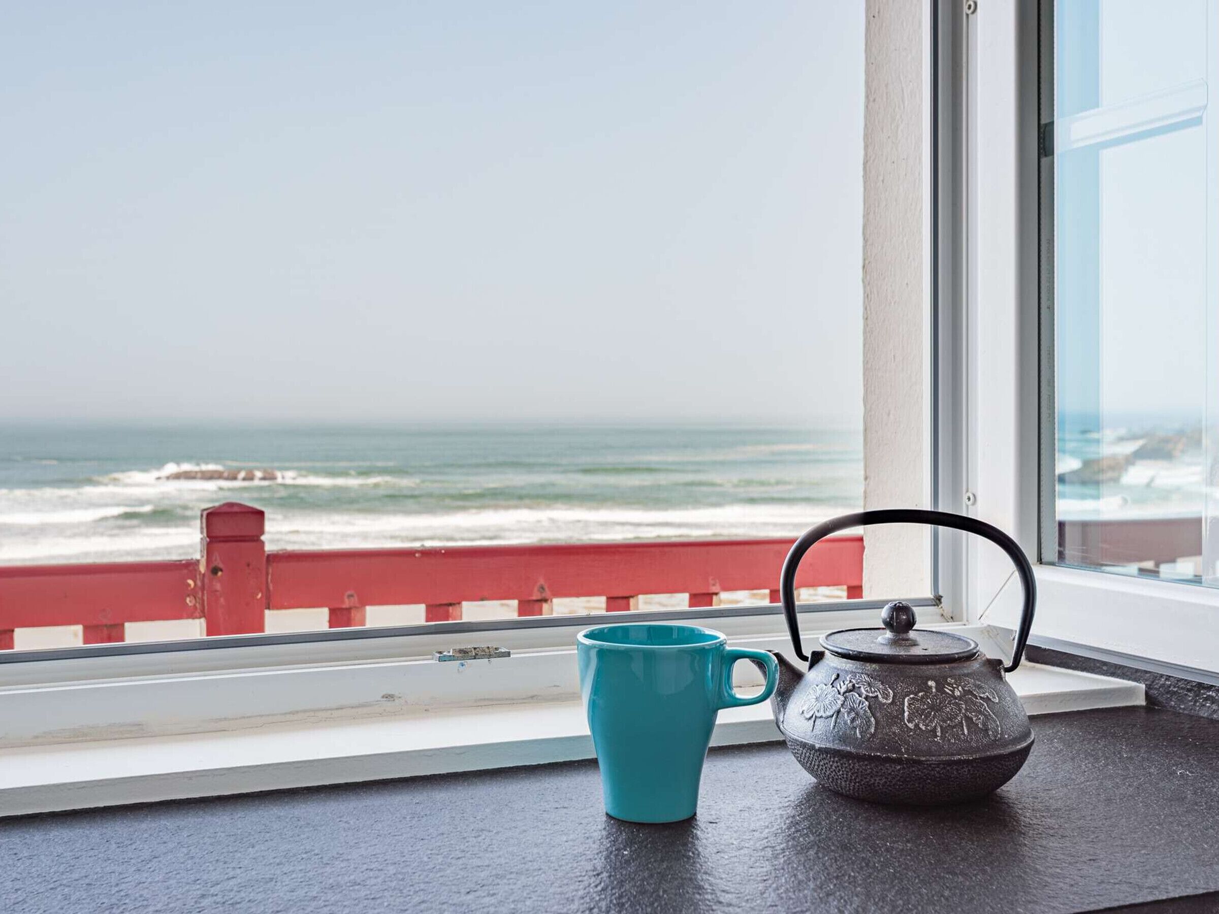Una ventana con vista al océano, barandilla roja, taza turquesa y tetera negra sobre el alféizar.