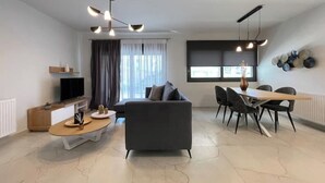 Ein modernes Wohnzimmer mit einer Couch, einem Esstisch und Stühlen, einem Fernseher und einem großen Fenster mit Vorhängen.