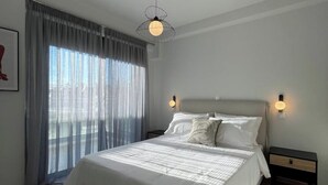 Un dormitorio moderno con una cama grande, dos mesitas de noche, una lámpara colgante y vistas a través de cortinas translúcidas.